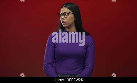 Woman in purple top wearing glasses crossing arms with serious expression in studio with red background; defiance. Stock Photo