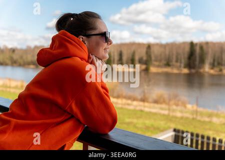 Caucasian Woman In Orange Hoodie Daydreaming Balcony, Hand Near Face, Overlooking Calm River And Distant Forest, Soft Clouds And Gentle Breeze Stock Photo