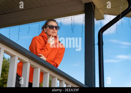 Caucasian Woman In Orange Hoodie Posing Confidently On Porch With Crossed Arms, Sunglasses Framing Face, Bold Expression Against Lake And Treeline Stock Photo