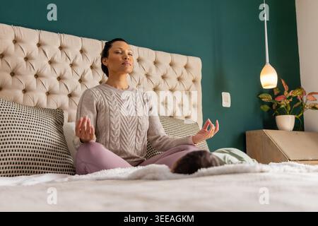 Mid adult woman meditating on bed in sweater and leggings with infant near headboard, lamp Stock Photo