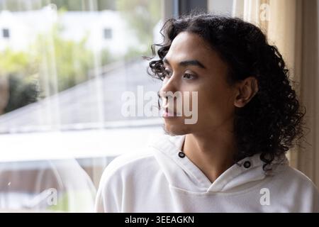 Non-binary person gazing toward right through large window at home, wearing white hoodie Stock Photo