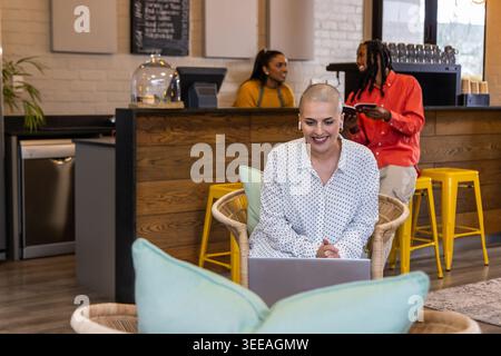 Diverse coworkers chatting by counter while woman sitting in rattan chair working on laptop in cafe Stock Photo