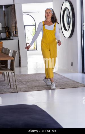 Middle-aged woman walking across bright modern interior at home, wearing yellow overalls Stock Photo