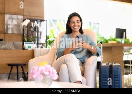 Smiling adult woman guest using smartphone at hotel lobby Stock Photo