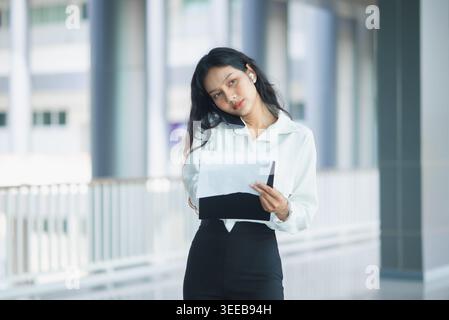 Busy businesswoman holds documents and talks on a smartphone hands-free while walking through a modern office corridor, showing multitasking, mobile w Stock Photo