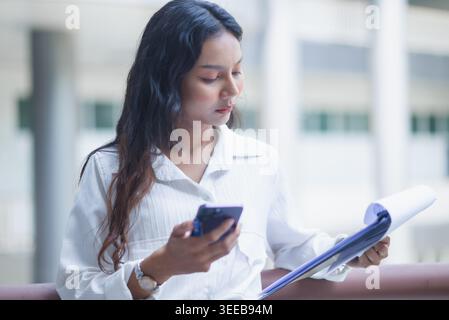 Confident businesswoman with wireless earbuds looks at the camera while writing on financial reports in a modern office stairwell, ideal for corporate Stock Photo