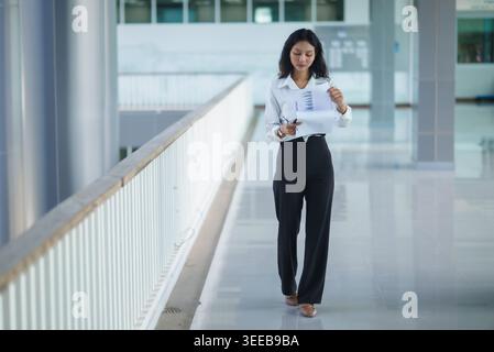 Thoughtful businesswoman holds a pen and smartphone while reviewing documents near an office railing, representing decision making, business planning, Stock Photo