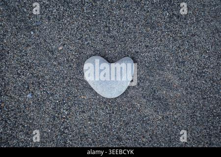 Smooth heart-shaped stone on a grainy sand - a natural symbol of love. Perfect for Valentine’s Day themes, wellness branding or environmental concepts Stock Photo