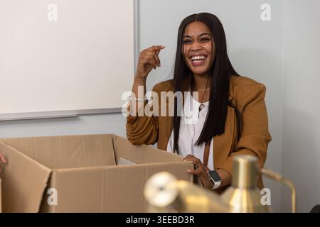 Mid adult African American woman in blazer laughing, gesturing and packing cardboard box at office Stock Photo