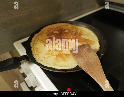 Pancake frying in a black pan on a ceramic hob, with a wooden spatula ready for flipping. Preparing homemade breakfast food Stock Photo