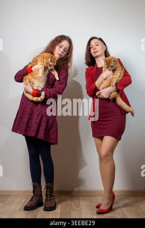 Two women stand against a neutral wall holding ginger tabby cats, wearing coordinated burgundy outfits and contrasting footwear. Stock Photo