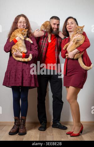 Smiling family of three holding orange tabby cats against a neutral wall, casual coordinated outfits, indoor studio portrait. Stock Photo