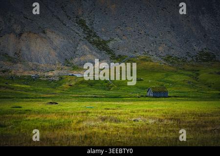Small rural house in green Icelandic valley beneath rocky mountain slope illuminated by sunlight. Stock Photo