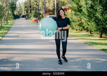 Fitness woman exercises with fitness ball outdoor walks on road near forest has healthy lifestyle dressed in black active wear, has happy expression, stays in good physical shape. Fitness concept Stock Photo