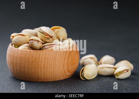 Close up, macro of salted pistachio nuts in wooden bowl with seeds scattered on textured dark gray background. Healthy snack food photography, organic Stock Photo