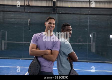 Posing two men wearing athletic T-shirts and dark shorts on blue padel court, holding padel rackets Stock Photo
