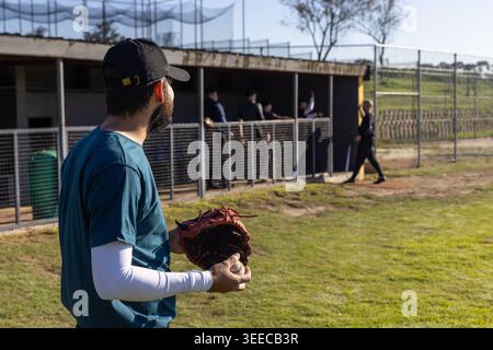 Male players leaning on dugout railing while bearded man wearing black cap holding ball and glove Stock Photo