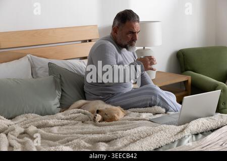 Senior man sitting at home on bed in pajamas holding mug and using laptop with dog Stock Photo