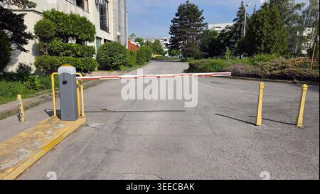 A motorized red and white gate arm blocks vehicle access on a grey paved street lined with green shrubs and yellow bollards leading toward urban apart Stock Photo