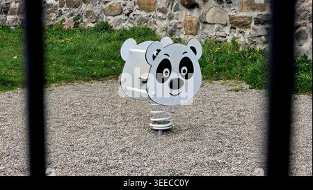 A white and black panda-themed spring rocker sits on a pebble-covered playground surface next to an old stone wall, partially framed by vertical black Stock Photo