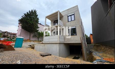 A three-story raw concrete house stands under construction featuring integrated window frames, an open garage level, and tiered balconies at a muddy Stock Photo