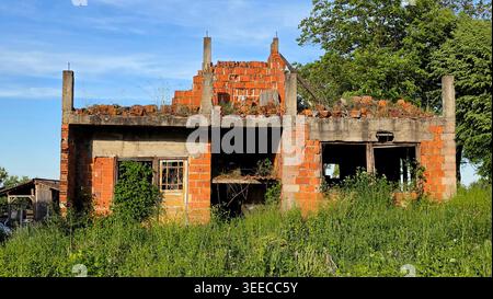 An abandoned two-story brick structure stands unfinished and roofless with tall green grass and weeds reclaiming the construction site under a clear Stock Photo