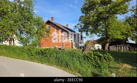 A two-story residential building made of red hollow clay blocks stands partially finished among lush green trees and a grassy field along a curving Stock Photo
