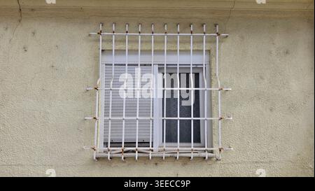 A residential window is protected by a white iron security grate and features partially closed plastic roller shutters set into a weathered, cracked Stock Photo