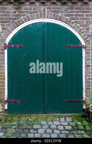 A rustic green wooden double door with an arched top is set within a traditional red brick wall. Featuring large metal hinges and a cobblestone foregr Stock Photo