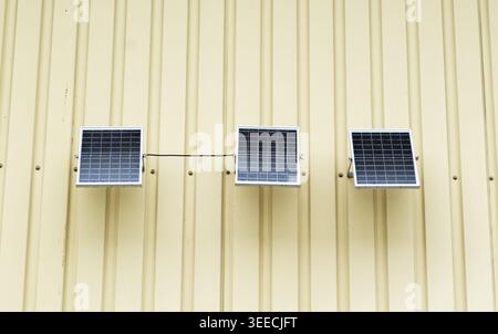 Three small solar panels mounted on a yellow corrugated metal wall for renewable energy generation Stock Photo