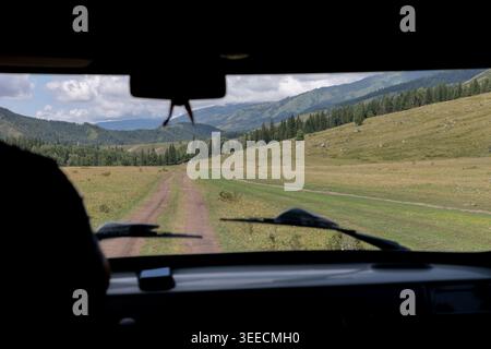 View from inside a vehicle driving along a rural dirt road through Ak-Kem Valley, Altai Republic, Russia, with green meadows, pine forest and mountain Stock Photo