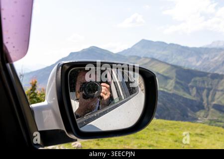 Travel scene in Dagestan, Russia: photographer taking a picture reflected in a car side mirror with green Caucasus mountain landscape in the backgroun Stock Photo