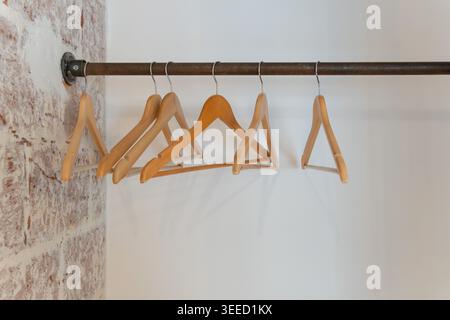 Wooden clothes hangers hang on metal rail against white wall, exposed brick, creating minimal interior detail with clean boutique mood. Retail design Stock Photo