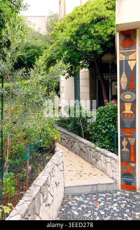 Decorative mosaic pathway leading to lush green garden of traditional Mediterranean villa in Nice. Colorful ceramic tile wall art with vase motifs Stock Photo