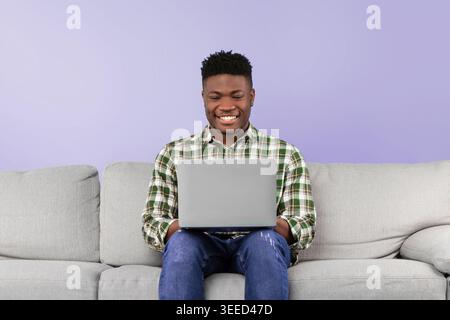 Smiling Man Using Laptop While Sitting on Gray Sofa in a Modern Indoor Setting Stock Photo