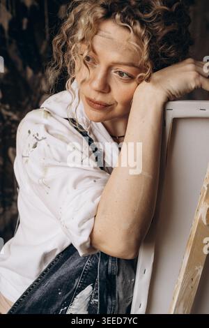 A young female artist pondering an idea for a future painting leaning on a canvas while standing in her creative studio. Painter portrait Stock Photo