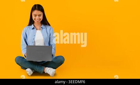 Young Woman Sits on Floor Using Laptop Against Yellow Background in Well-Lit Space Stock Photo