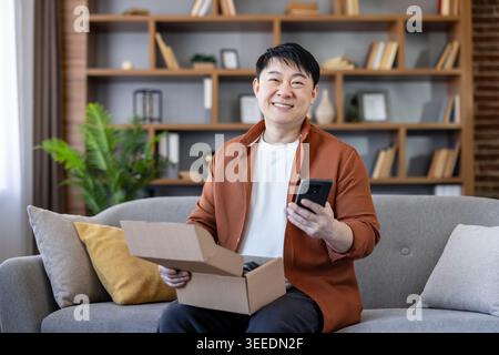 Smiling asian man sitting on a couch, holding an open cardboard box with newly received goods and using his smartphone, happy with his online shopping delivery experience at home Stock Photo