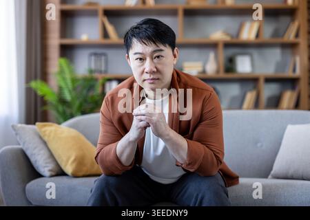 Mature asian man sits on a sofa with clasped hands, staring at the camera in quiet contemplation, conveying worry, solitude and thoughtful concern at home in the living room Stock Photo