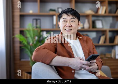 Asian man smiling, sitting comfortably on a sofa at home, using his smartphone for communication, entertainment, and modern connection in a relaxed informal setting Stock Photo