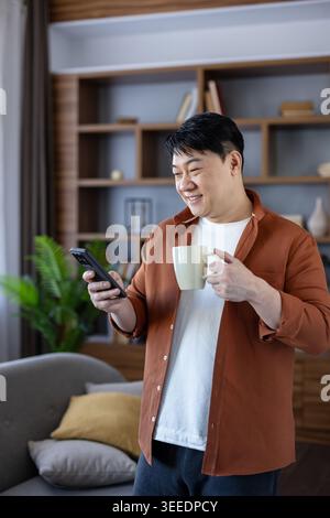Smiling asian man standing at home, holding a coffee mug and using his smartphone for communication, enjoying leisure time during a morning break in his living room Stock Photo