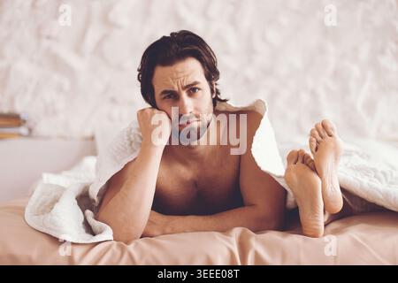Man Sitting on Bed Looking Thoughtful Under a Blanket in a Simple Room With Neutral Colors Stock Photo