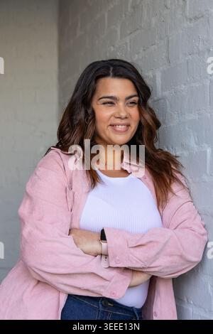 Woman leaning against painted brick wall in studio, wearing pink overshirt and smartwatch Stock Photo