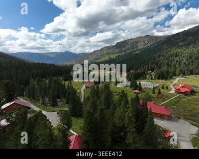A serene mountain valley features red-roofed wooden cabins surrounded by tall evergreen trees under a partly cloudy blue sky. Stock Photo