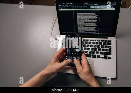 Top view of programmer hands holding smartphone and typing, viewing text messages while working on laptop with code displayed on screen, managing mult Stock Photo