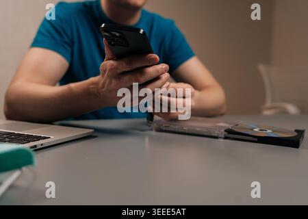 Cropped shot of man connecting smartphone to external optical disc drive on a desk beside laptop, showing a media disc and glowing blue usb ports for Stock Photo
