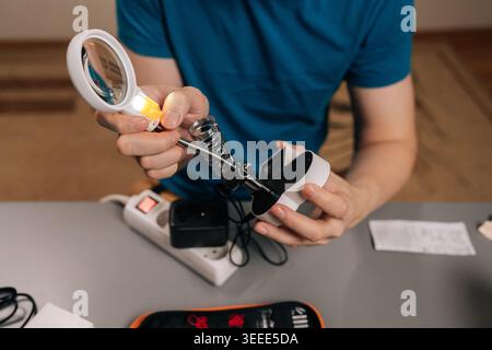Close-up cropped shot of technician working on electronics using a soldering iron and a magnifying lamp, carefully assembling or repairing a circuit b Stock Photo