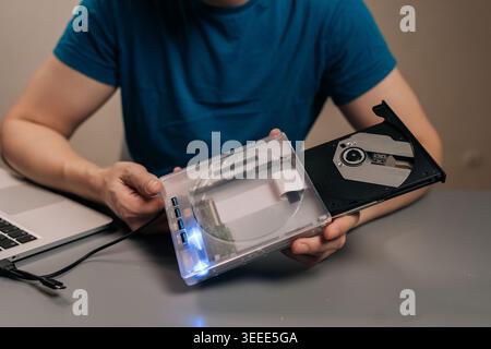 Close-up hand of person holding transparent external optical drive with open disc tray connected to laptop on desk, demonstrating portable dvd cd read Stock Photo