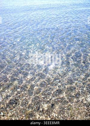 Clean transparent water on shallow sandy seashore with shells with shells on sunny summer day. Sun glare on surface of water. Small waves on sea coast. Nature backdrop. Natural background Stock Photo