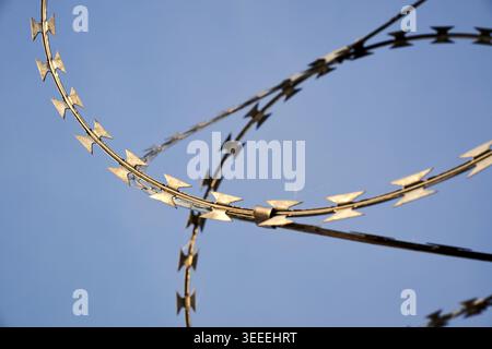 Close-up shot of gleaming concertina razor wire coils forming a formidable security barrier set against a bright, cloudless blue sky. The sharp metall Stock Photo
